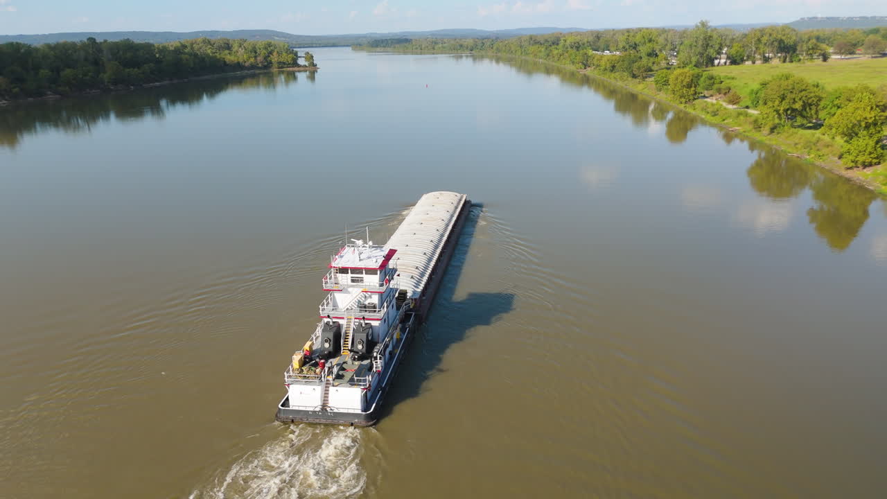 Drone pullback as barge floats down Arkansas river, trees on riverbanks and bright sunlight overhead