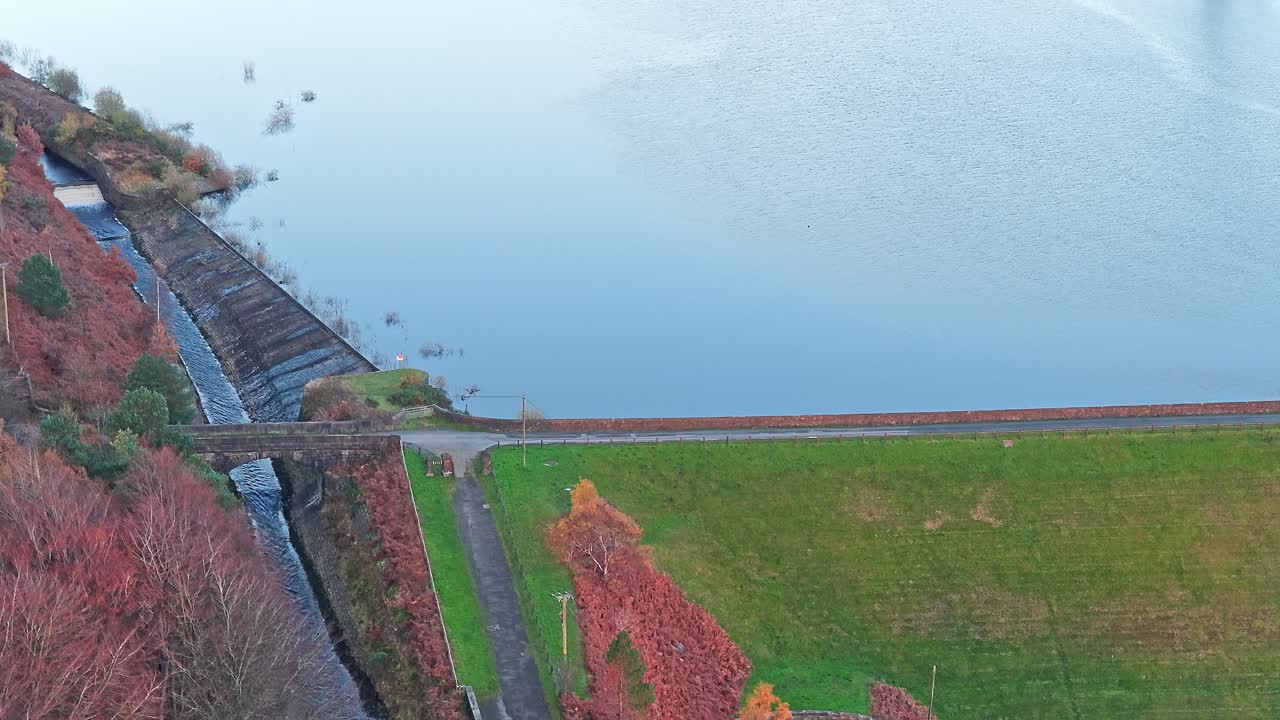 Overhead details stepped concrete spillway, road across the grassy Woodhead Embankment dam, and tranquil reservoir water at the head of the Longdendale Valley, on adjacent woodland in Pennines