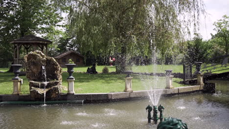 Tranquilo-Jardín-Con-Una-Fuente,-Un-Mirador-Y-Una-Exuberante-Vegetación,-Con-Salpicaduras-De-Agua-Y-Un-Puente-De-Madera,-Que-Crean-Un-Ambiente-Sereno-Al-Aire-Libre