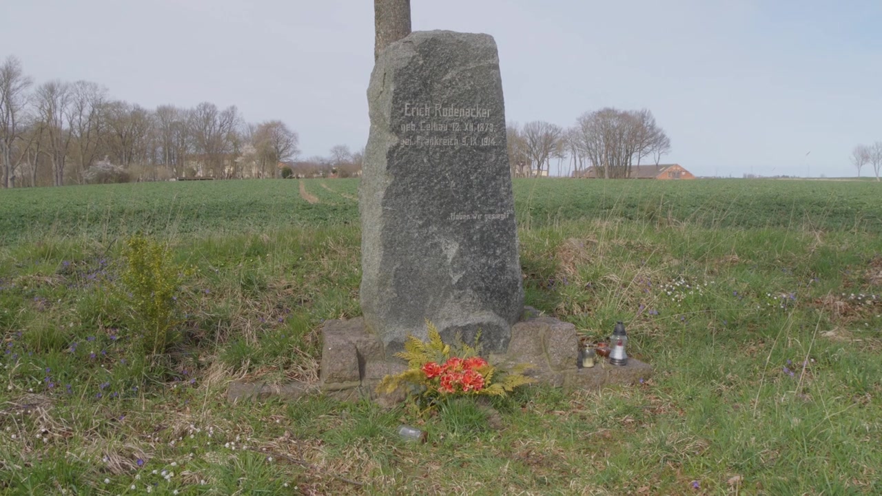 Storico monumento funebre della famiglia Rodenacker a Celbowo, Polonia, con un obelisco di pietra eroso, candele e fiori accanto a campi rurali aperti.