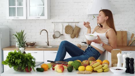 happy girl eats salad in a white kitchen near a table with fruits and vegetables