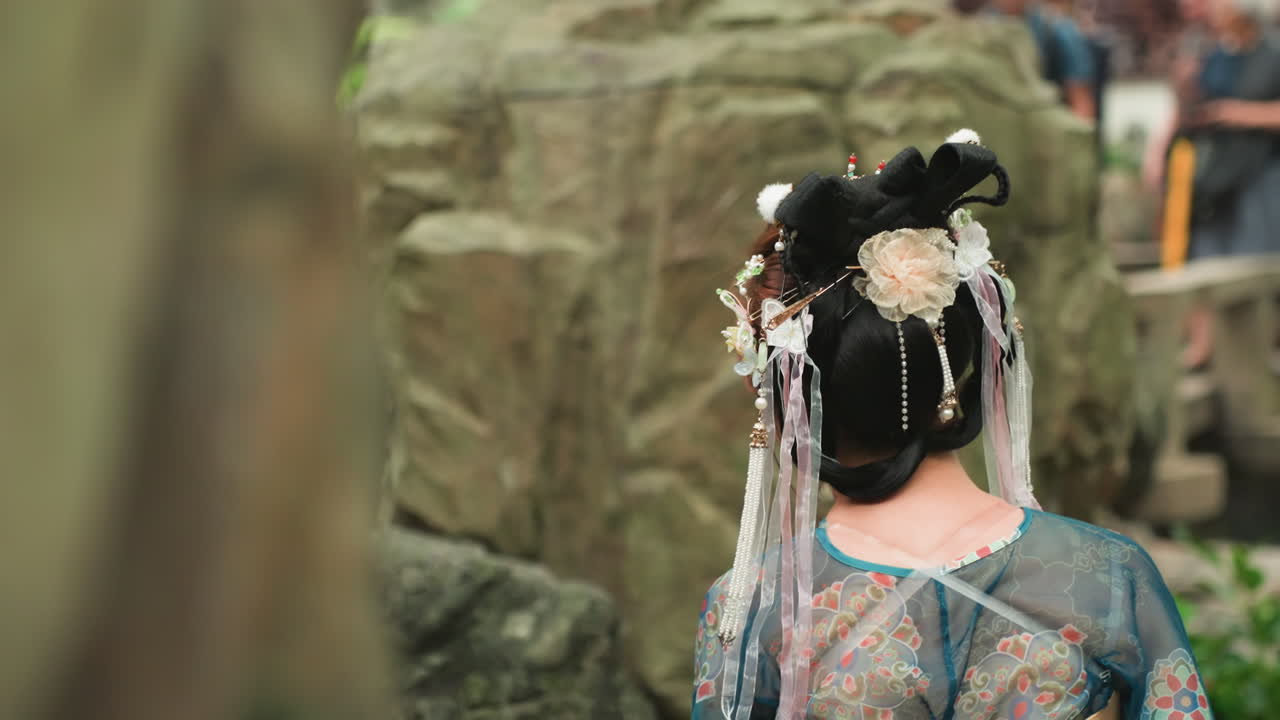 Vista posteriore di una performer asiatica con ornamenti per capelli sontuosi e un costume di seta ricamato, in piedi accanto a una formazione rocciosa in un giardino lussureggiante, nappe di perline che le scendono tra i capelli, spettatori sfocati sullo sfondo.
