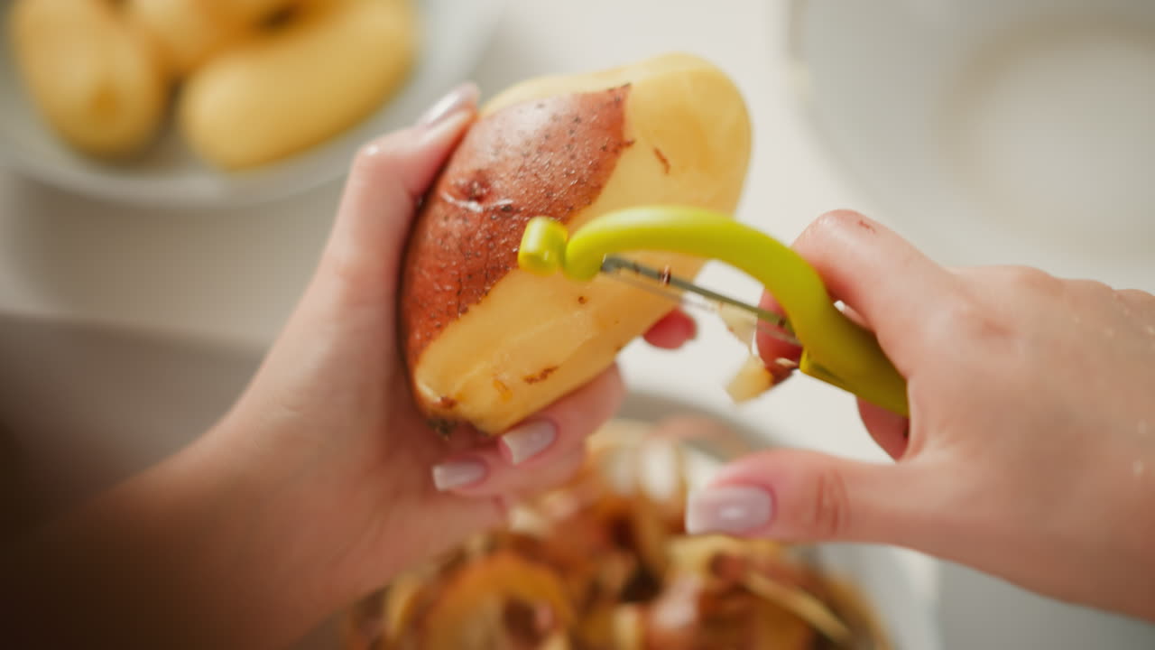 Close up of person peeling potato with green peeler tool, skin partially removed as peeled potatoes blur in background, creating warm and detailed representation of kitchen preparation activity