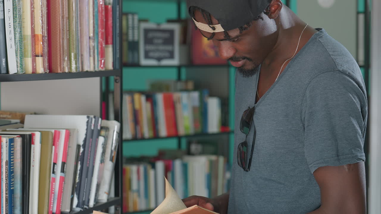 Young man in casual clothing and backward cap standing near bookshelves in library, holding open book and reading attentively, surrounded by colorful spines
