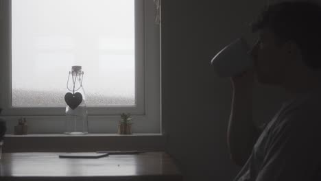 person drinks from mug and looks out window as it is raining heavily and about to storm