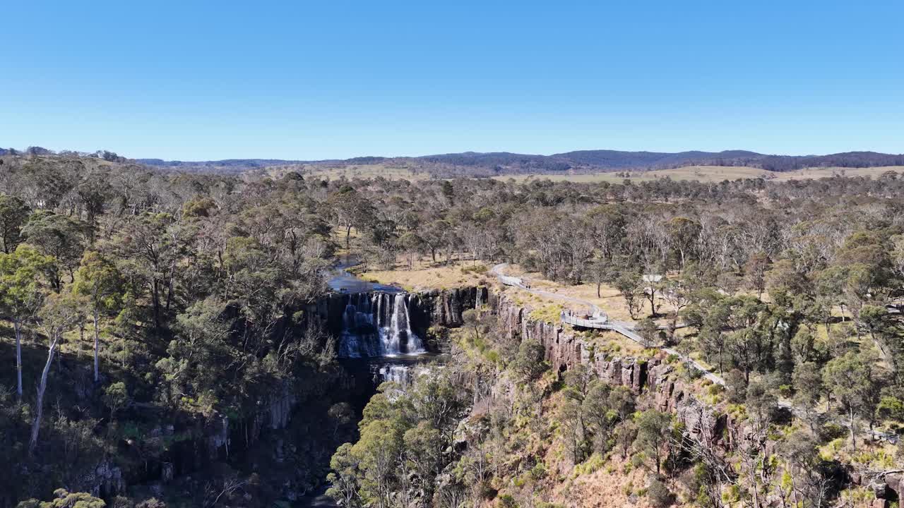 Drone camera glides over Ebor Waterfall and surrounding forested gorge in bright daylight, revealing rugged cliffs, flowing water, and expansive New England landscape
