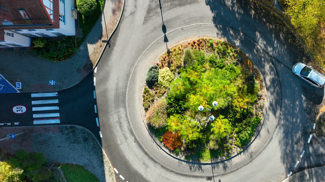 Top-down drone shot of a circular roundabout in Sopot, Poland, featuring a lush green island with colorful plants and surrounding urban streets