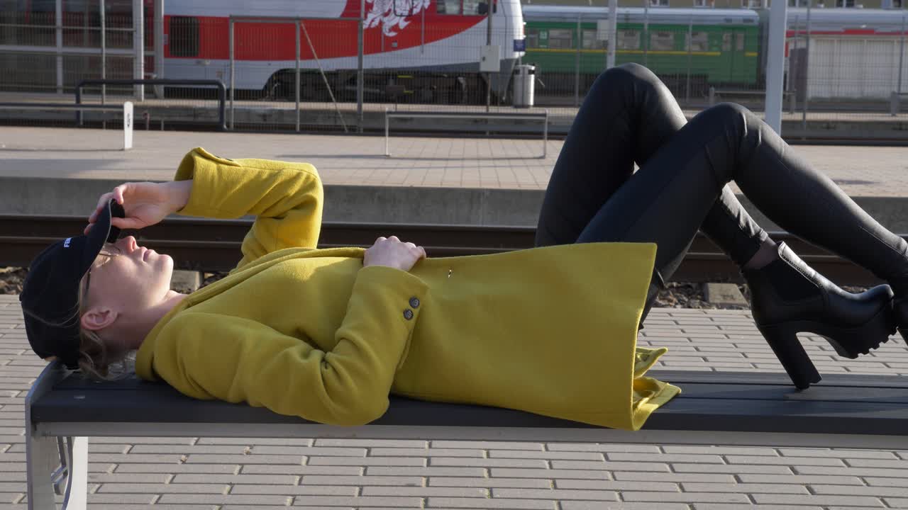 A young beautiful woman in high heels rests on a  bench on a platform while waits  for a train.