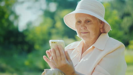 An-Elderly-Woman-In-Summer-Clothes-And-A-Hat-Uses-A-Smartphone-Relax-In-The-Summer-Park-4K-Video