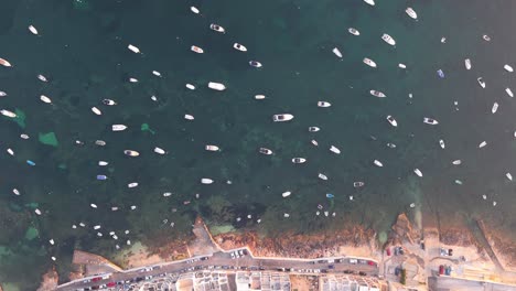 a drone view of a mediterranean coastal town showing tightly packed buildings, a seaside road, and numerous boats anchored in the turquoise water