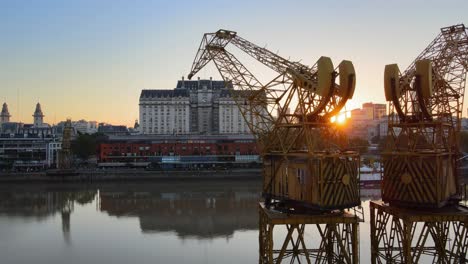 Toma-Aérea-De-La-Derecha-Del-Carro-De-La-Puesta-De-Sol-Detrás-De-Las-Viejas-Grúas-Portuarias-En-El-Paseo-Marítimo-De-Puerto-Madero,-Buenos-Aires