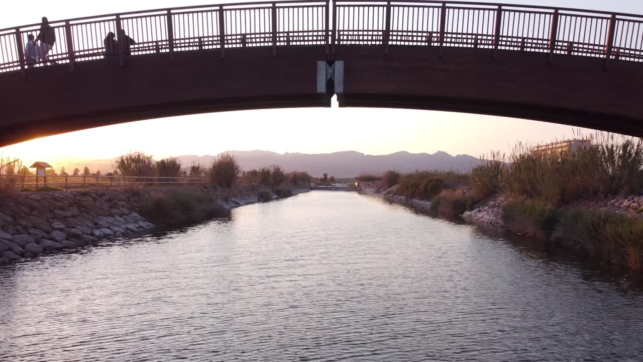 A drone flying directly under the middle of an arch bridge at sunset with people stood and walking across the bridge. Moving towards with mountains in the distance