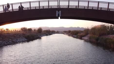 a drone flying directly under the middle of an arch bridge at sunset with people stood and walking across the bridge. moving towards with mountains in the distance