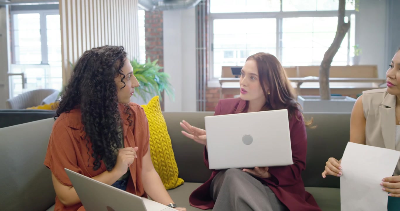 Team femminile che lavora in una lounge, quella al centro mostra un laptop, quella a sinistra indica uno schermo, quella a destra tiene dei documenti.