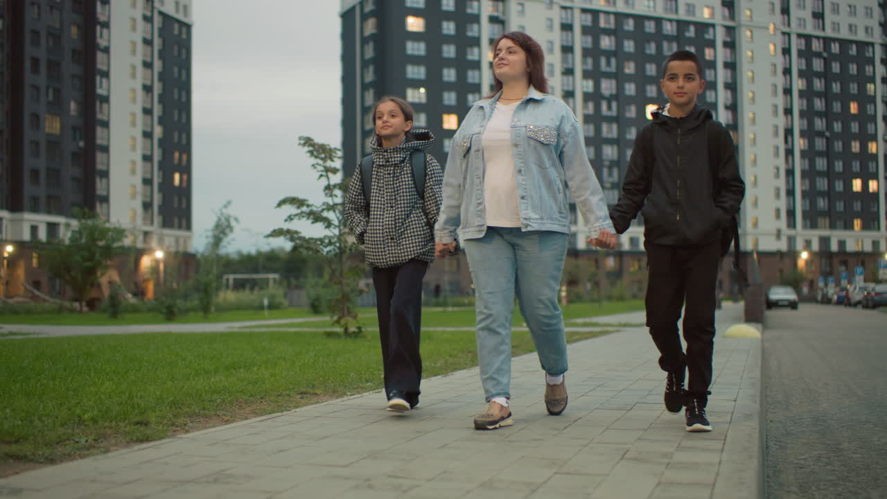 Una mujer camina de la mano con dos niños por un sendero pavimentado en una zona residencial moderna durante la tarde, rodeada de apartamentos de gran altura y césped verde, expresando unidad, confianza y calidez en la vida urbana cotidiana.