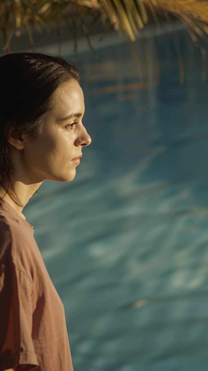Vídeo vertical: Mulher de pé com blusa malva fazendo movimentos com a cabeça enquanto a luz do sol atinge a piscina, espaço para cópia