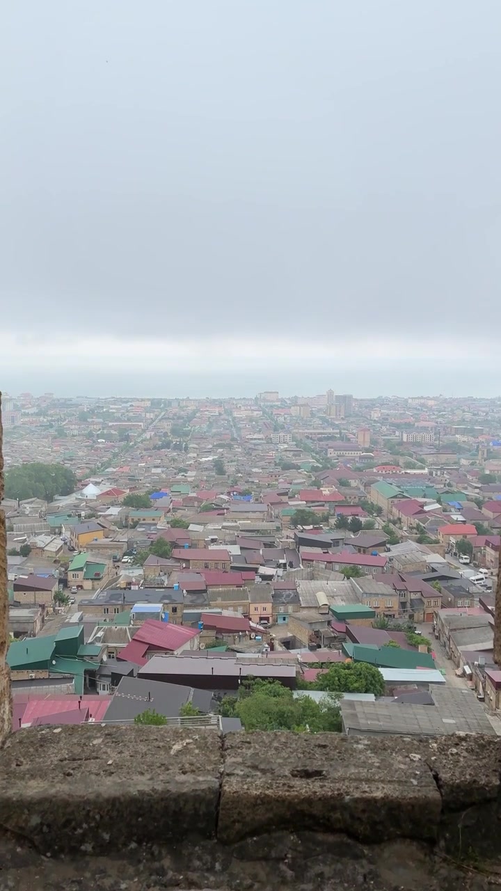 Sebuah kamera bergerak melalui bingkai jendela batu kuno, menyingkap pemandangan panorama kota Derbent dengan atap-atap berwarna-warni di bawah langit mendung