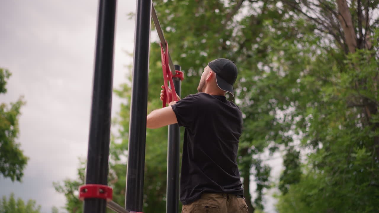 Male Examines Resistance Band, Man Adjusting Resistance Strap Beneath Overcast Sky At Park, Person Carefully Inspects Resistance Band Attached To Metal Frame In Park Under Cloudy Weather