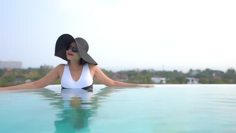 French-lady-in-Sunhat-and-Monokini-style-Swimsuit-Relaxing-Inside-Rooftop-Infinity-Pool-with-Cityscape-on-background,-travel-vacation-concept