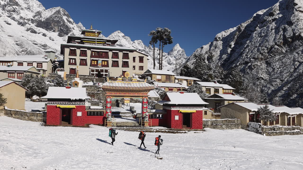 Three people walking past Tengboche Monastery carrying bags in the mountains of Nepal.