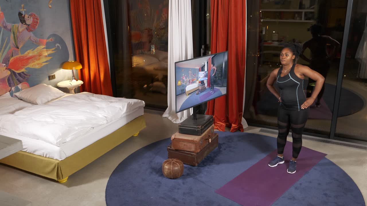 운동복을 입은 여성이 현대적인 침실에서 가정 운동을 하고, 저녁에 tv에서 운동 루틴을 따르고 있습니다.