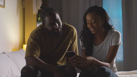 Young-Couple-Relaxing-At-Home-At-Night-In-Bedroom-Looking-At-Mobile-Phone-Together-4