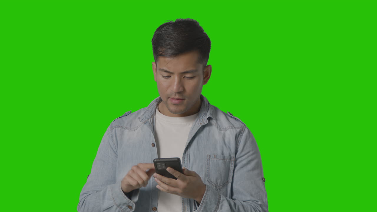 캐주얼한 옷을 입은 젊은 남자가 녹색 화면을 배경으로 휴대전화를 온라인으로 스와이프하는 스튜디오 샷 1