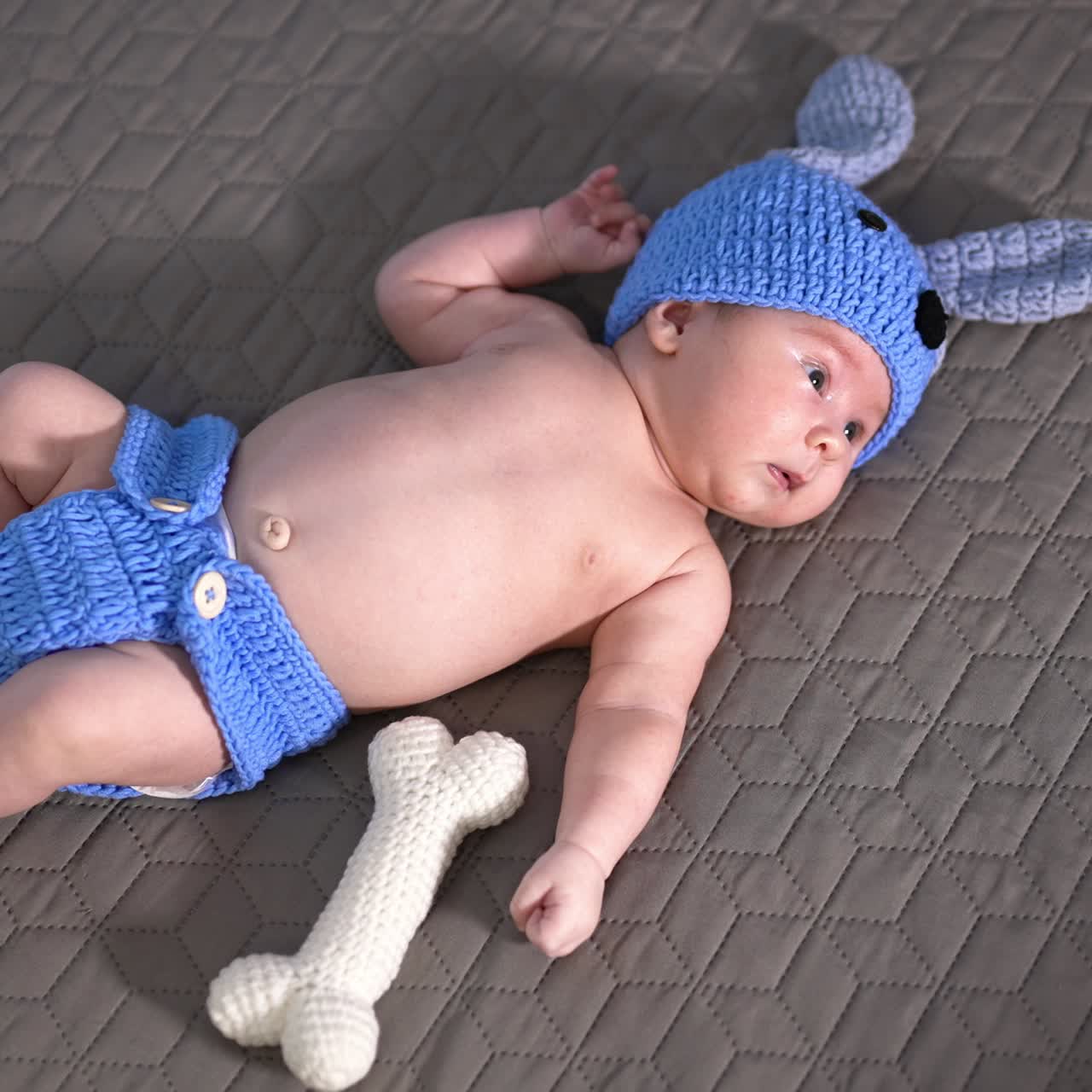 Newborn kid wearing funny blue knitted suit of a puppy. Toy bone next to the boy. Baby in a funny costume on the grey backdrop