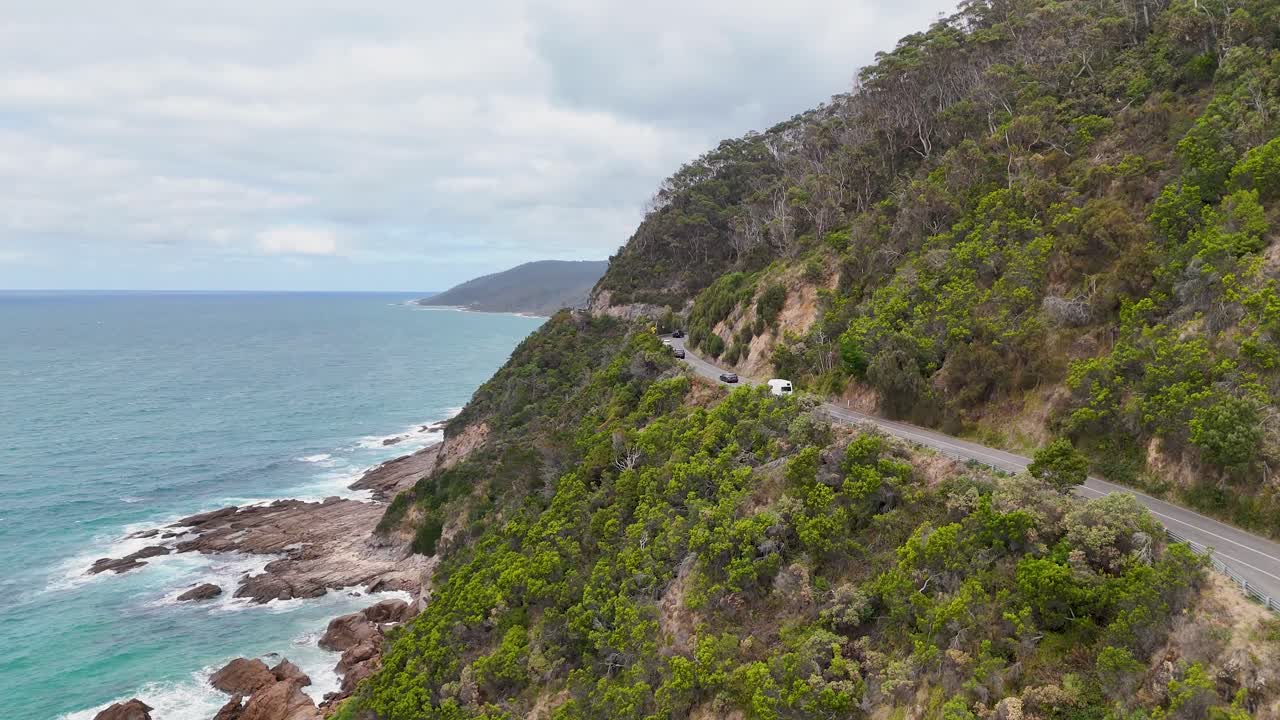 Aerial view of a car driving along the coastal Great Ocean Road in Lorne, Australia, surrounded by lush greenery and ocean views