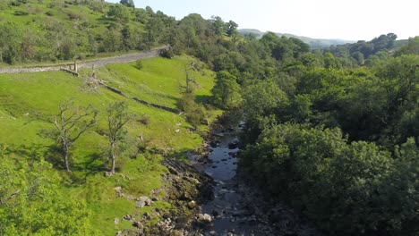 drone footage on a sunny summer day through a shallow river valley and exposed river bed, lined with grassy banks and trees with a country road on one side and railway on the other