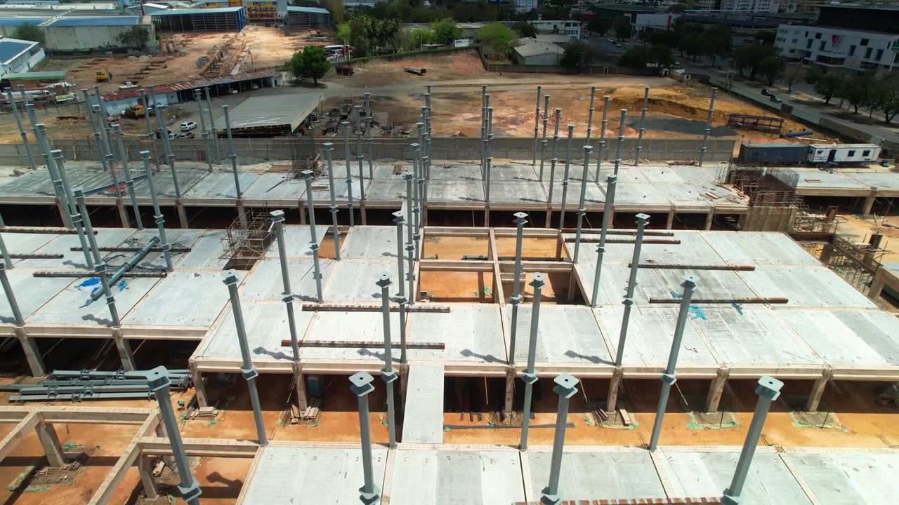 High-angle aerial drone shot flying backward and tilting up over a commercial building construction site, showing steel pillars, concrete floor slabs, and city skyline