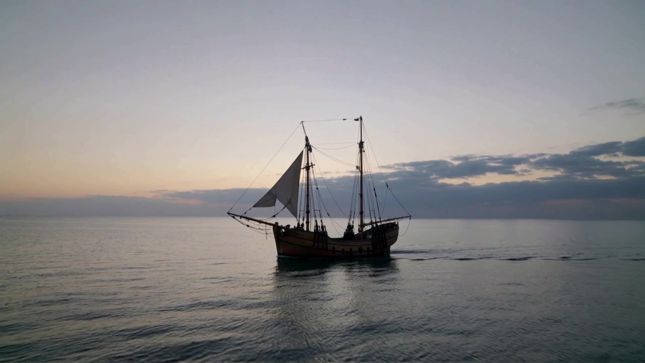 Vascello di legno a due alberi che naviga, avvicinandosi alla telecamera attraverso un mare calmo, con una leggera brezza che gonfia la vela di prua
