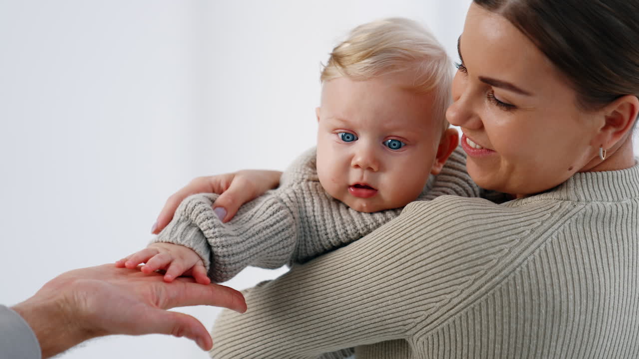 Blue-eyed blond infant in mom's hands. Cute baby touching and clapping male hand. White backdrop.