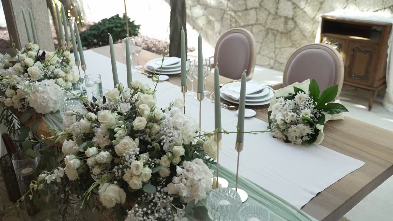 Elegant table arrangement with white floral centerpieces, tall candles, and stacked plates on a soft, neutral colored tablecloth