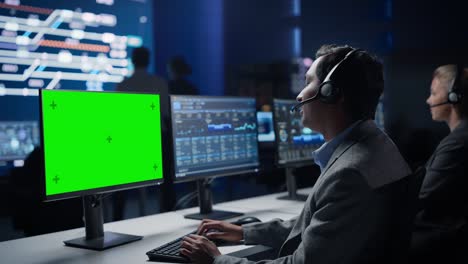 customer support specialist talks to client on headset works on green chroma key screen computer in big infrastructure control room. specialists use computers showing graphs. talk to investors