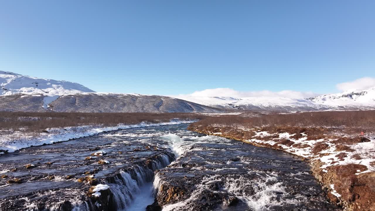 Panoramic aerial view of Brúarfoss waterfall, Iceland, featuring the distinct blue waters cutting through the snowy winter landscape.