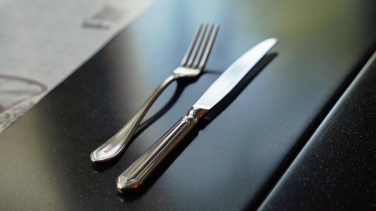 Gros plan cinématographique d'une fourchette et d'un couteau en argent poli sur une table sombre et mouchetée en plein jour. L'arrière-plan flou d'un menu de restaurant crée une faible profondeur de champ pour les publicités de restauration.