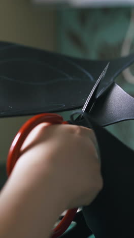 cu: skilled tailor cuts black leather cloth with scissors against blurry modern multicooker close view