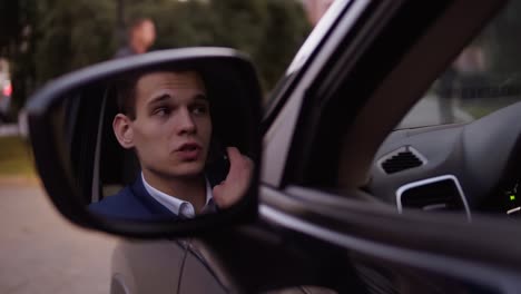 Handsome serious businessman in suit and white shirt talking by mobile phone in the car. Reflection in the car&#039;s door mirror