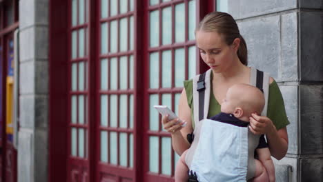 con su bebé en una mochila de canguro, una mujer joven documenta su viaje con un teléfono móvil. mientras camina, mira la pantalla del teléfono intermitentemente