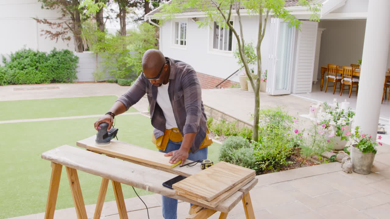Après serrage, un homme afro-américain portant une ceinture à outils ponce une planche sur une terrasse avec une ponceuse pour la finition