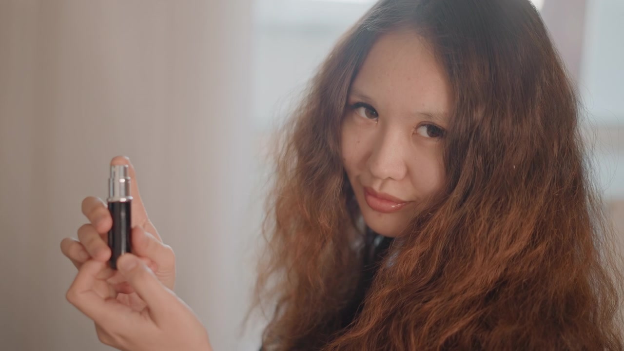 Giovane donna tiene un piccolo prodotto cilindrico vicino alla fotocamera con entrambe le mani, stabilendo un contatto visivo diretto e un leggero sorriso nella morbida luce della finestra per una presentazione commerciale calma.