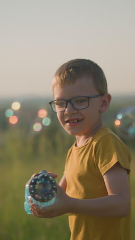 close-up of a young boy wearing a yellow shirt and shorts, with glasses, joyfully running through a sunny grassy field while holding a plastic bubble gun that releases a stream of bubbles