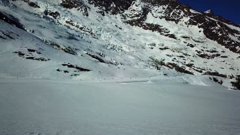 Drone-view-of-a-snow-plain-in-the-swiss-Alps,-chair-lift-and-skiers-in-the-background