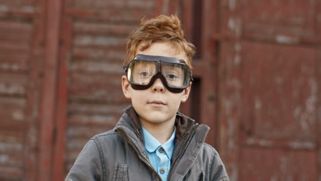 close up of a cute little boy with red hair and glasses putting on an aviator hat and smiling at the camera