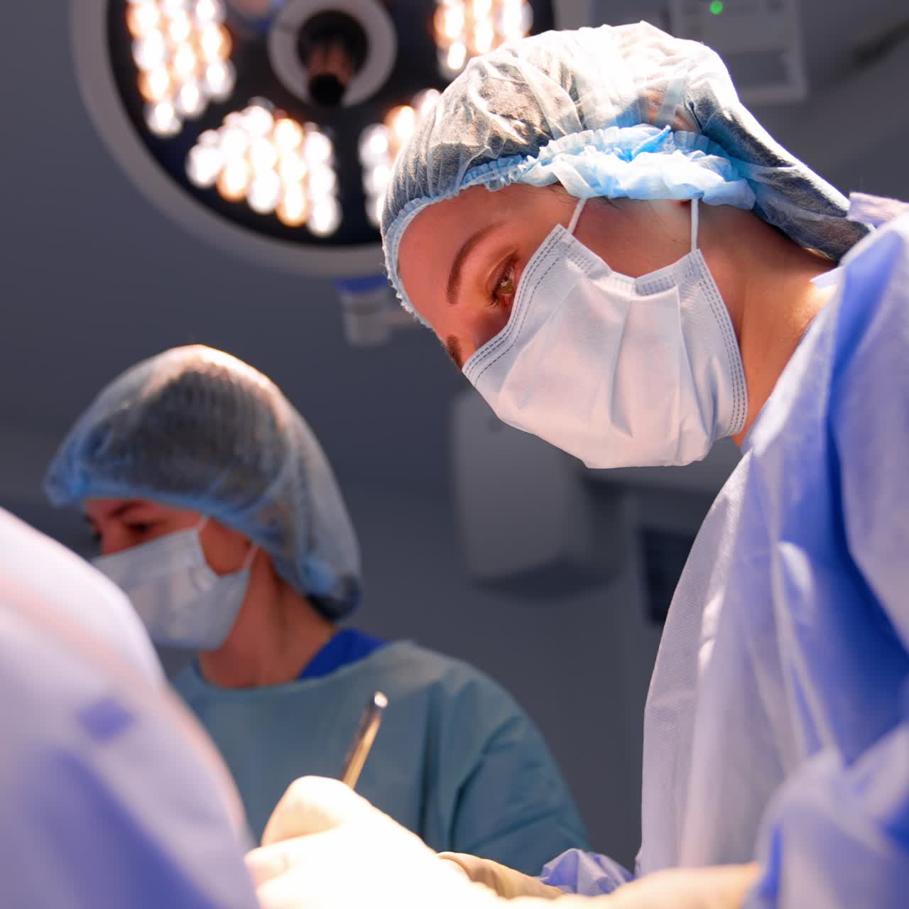 Una dottoressa con mascherina e cuffia è concentrata sul lavoro in sala operatoria. Angolazione dal basso sul chirurgo che esegue l'operazione