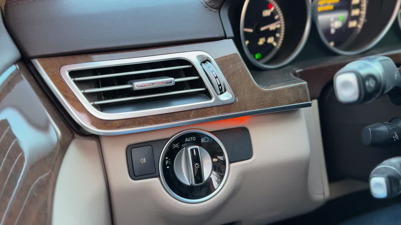 Un cruscotto di auto di lusso con una bocchetta dell'aria cromata, finiture in legno lucido e una manopola di controllo dei fari. La luminosa illuminazione naturale evidenzia la pelle pregiata e i dettagli interni