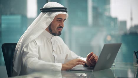 arab businessman in white traditional outfit sitting in office and working on laptop computer. business manager celebrates successful investment deal. saudi, emirati, arab businessman concept.