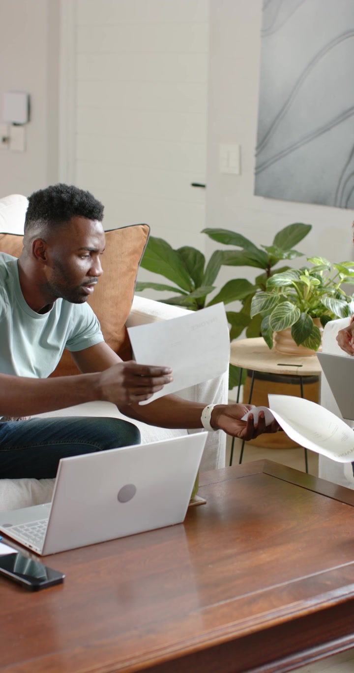 Vertical video: Diverse couple collaborating on couch man coming left handing papers by laptop