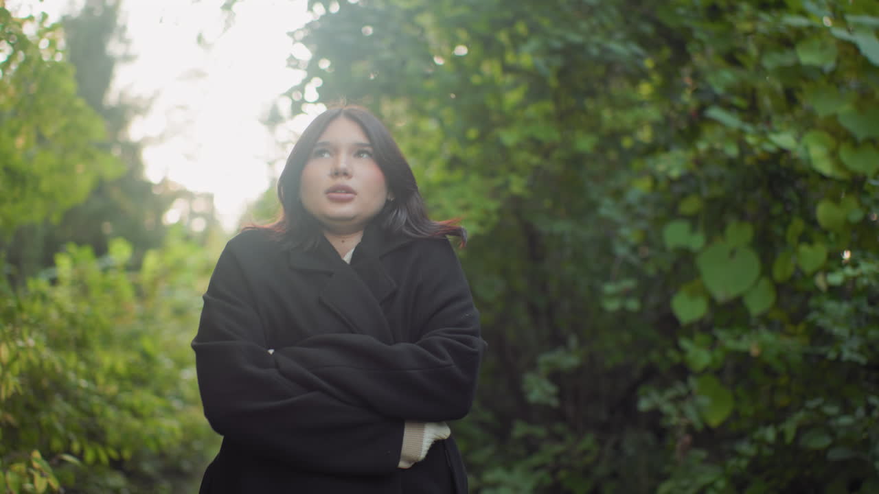 Serene lady adjusts coat while walking through green forest, breathing deeply, looking around with calm focused gaze, soft daylight on leaves, mindful stroll with warm sweater and black outerwear